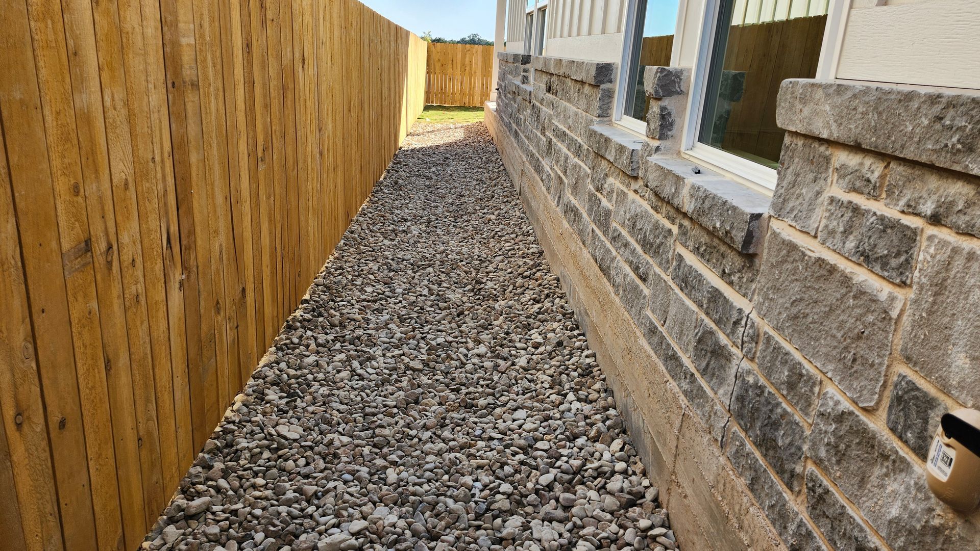 Gravel path between a wooden fence and a brick building with a window. Sunny day.