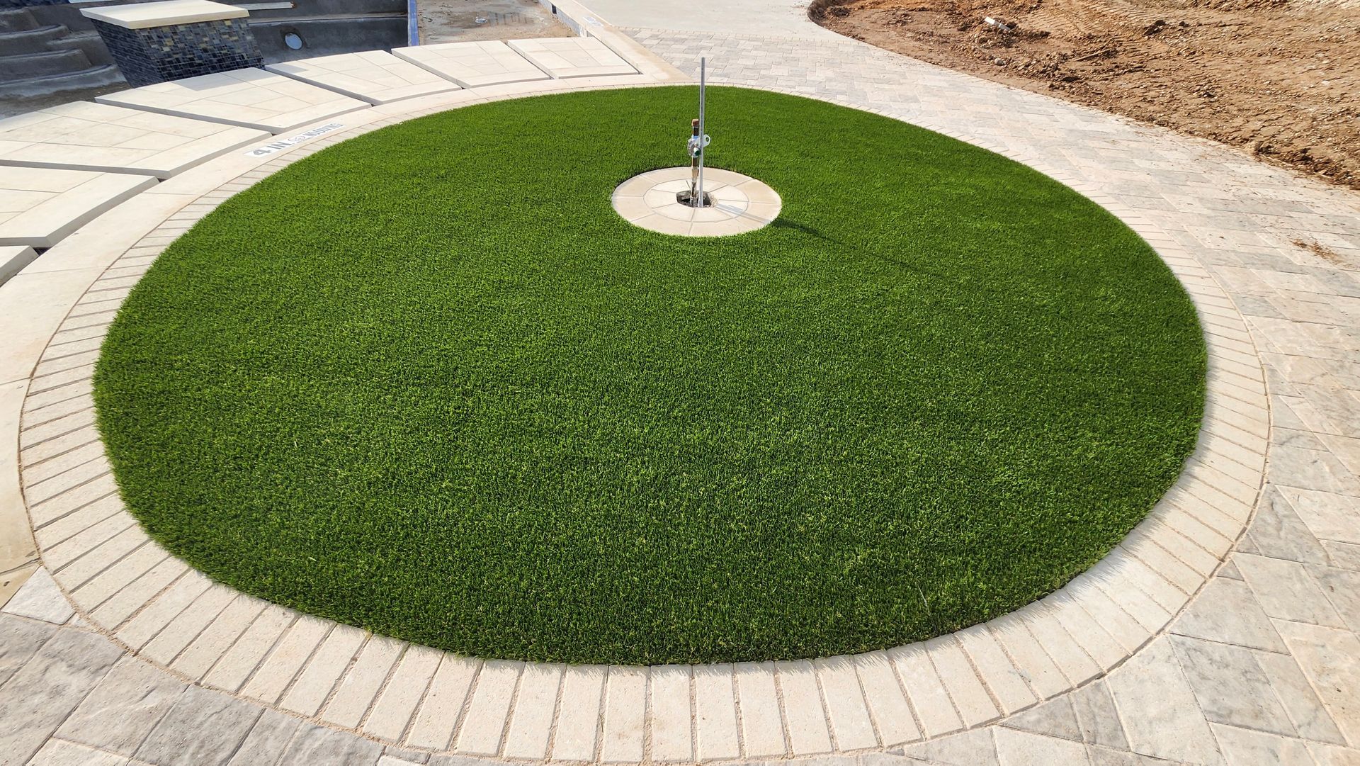Circular green lawn with a water feature, surrounded by tan brick and stone paving.
