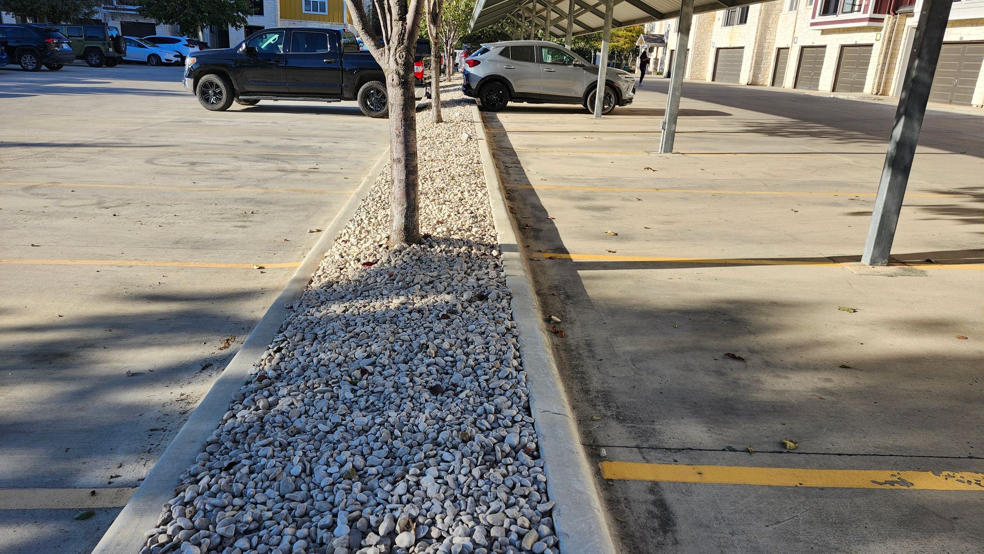 Parking lot with a gravel-filled planter between parking spaces. Cars and a metal shelter are visible.