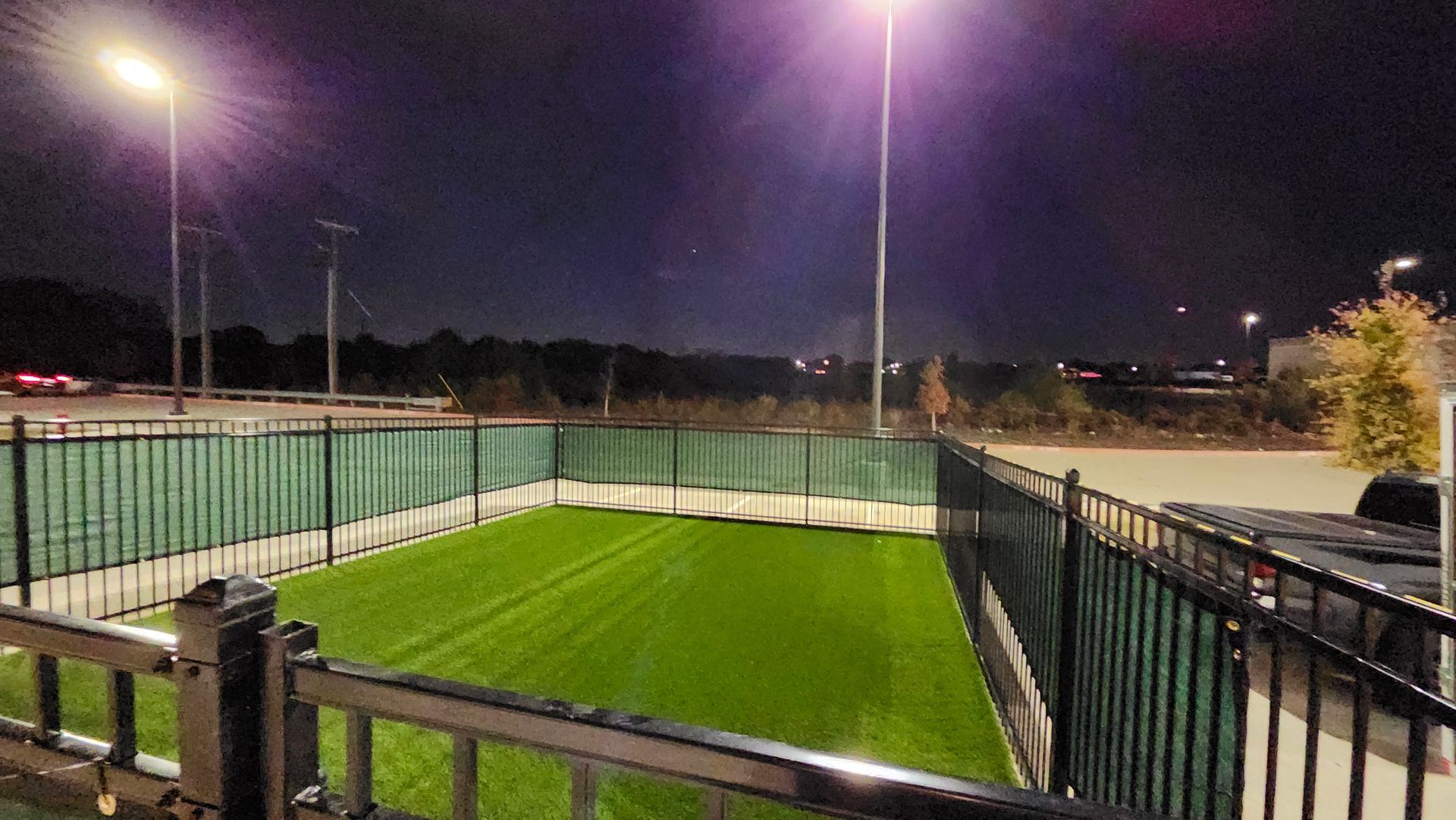 Nighttime view of a fenced, green artificial turf area under bright streetlights.