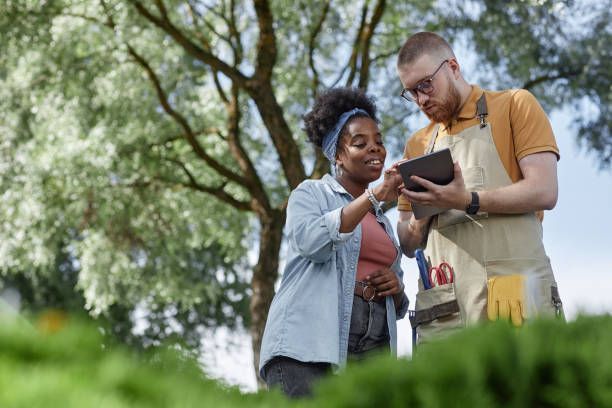 A person in a tan apron with gardening tools consults a tablet with another individual outdoors in a leafy, green setting.