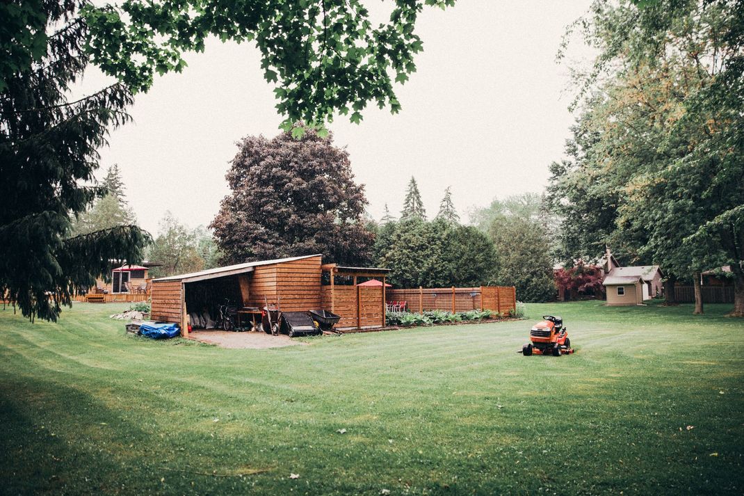 Lawn being mowed in a backyard with a wooden fence, shed, and trees. Cloudy day.