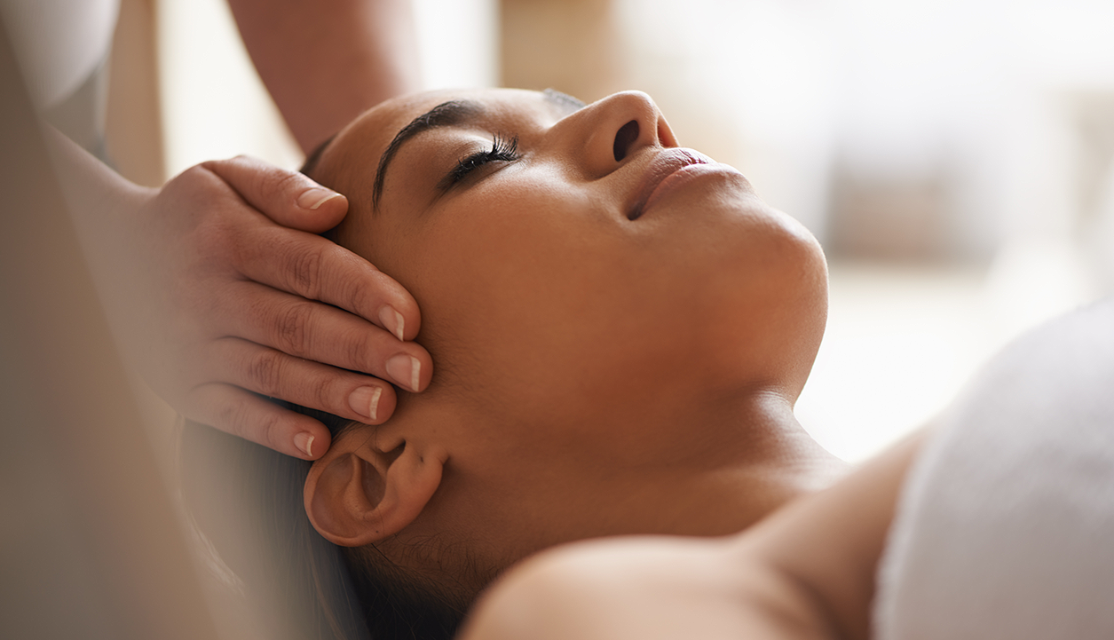 A beautiful woman on her back with a serene look on her face as someone's hands massages her temples.