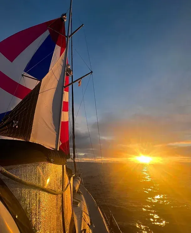 Sailboat with flag sailing on water at sunset. Sun reflects on water.