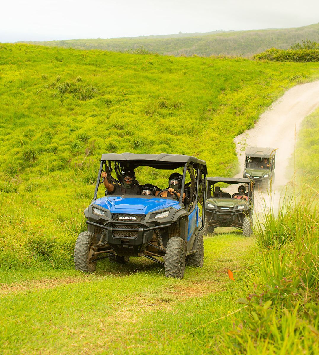 Off-road vehicles driving on a dirt path through a grassy hill. One blue vehicle leads the way.