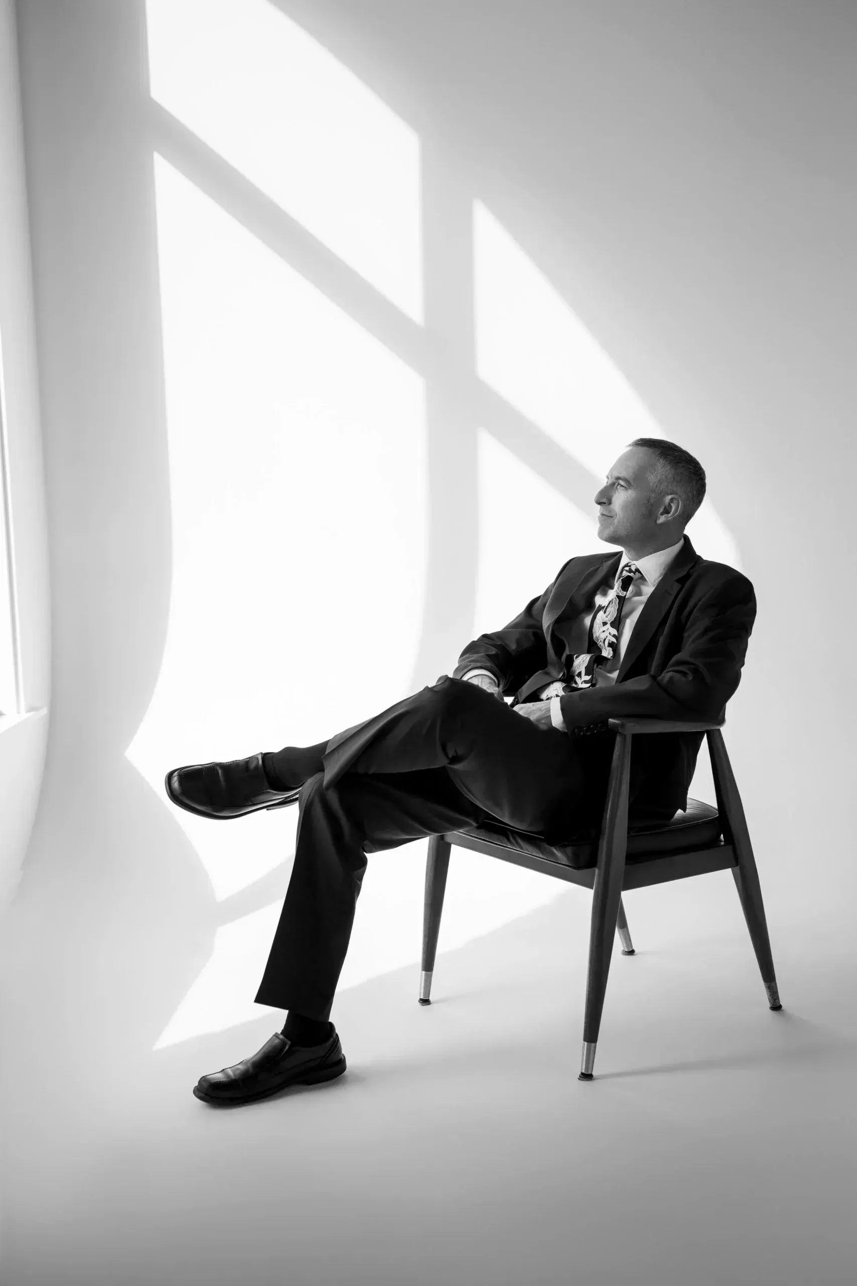 Man in suit sits in a chair, looking right, with sunlight shadows on the white wall.
