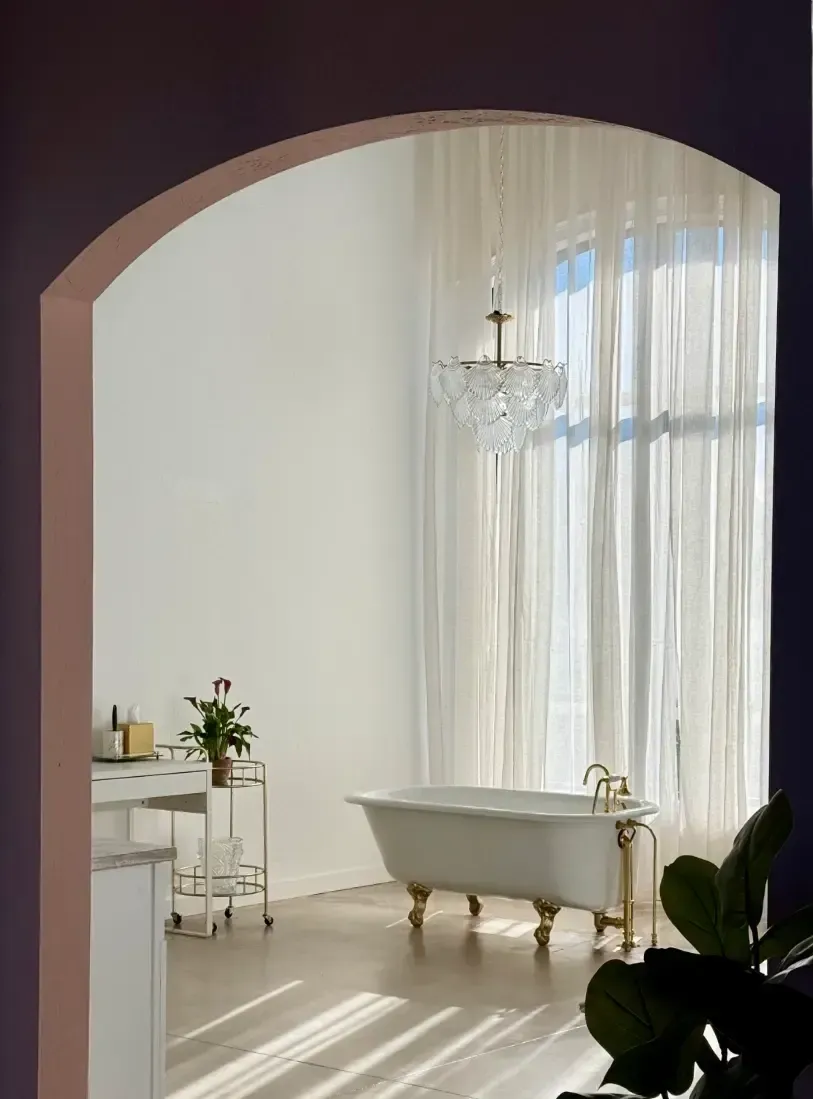 Archway view of a bright bathroom with a clawfoot tub, chandelier, and sheer curtains.