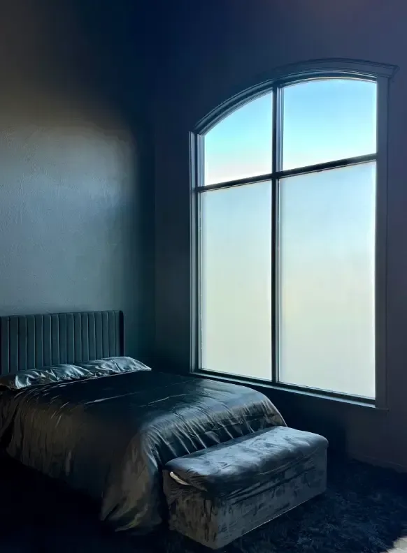 Bedroom with a tall arched window, bed, and bench.  Soft gray tones and diffused light.