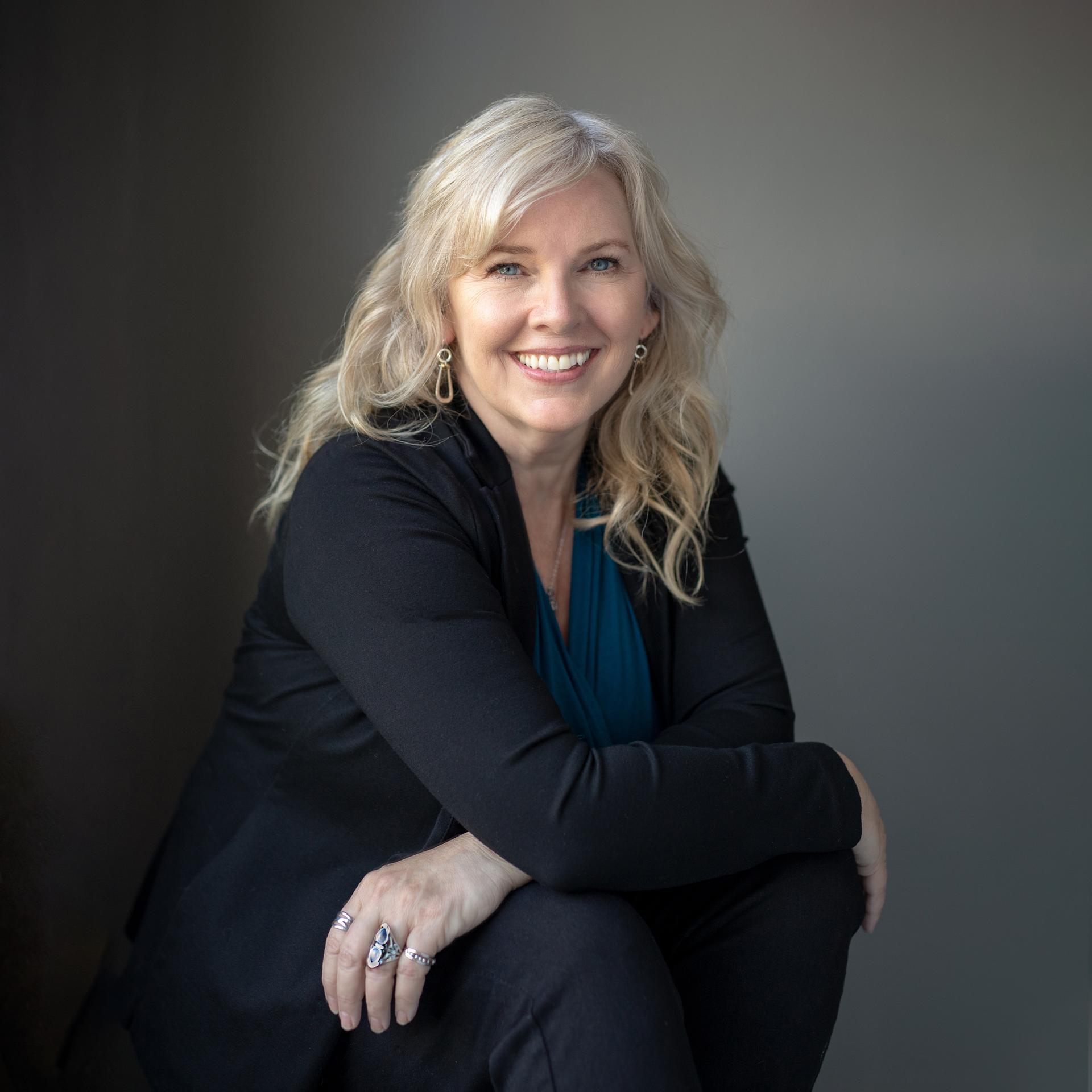Woman with blonde hair smiles, seated, leaning against a neutral background. She wears a black blazer, blue shirt, and jewelry.