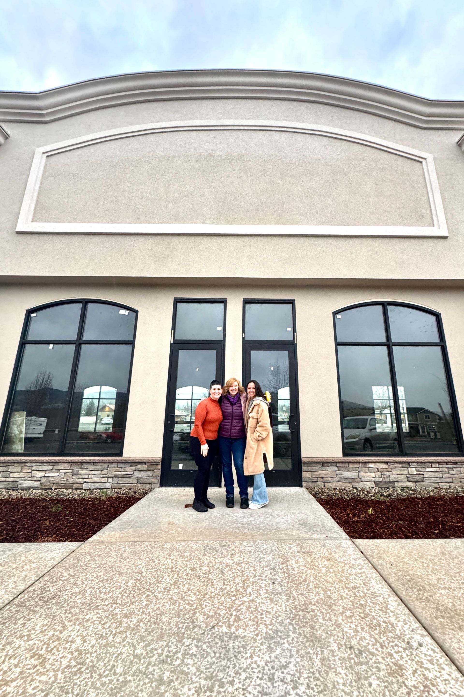 Three people stand in front of a tan commercial building with large windows. Cloudy sky overhead.