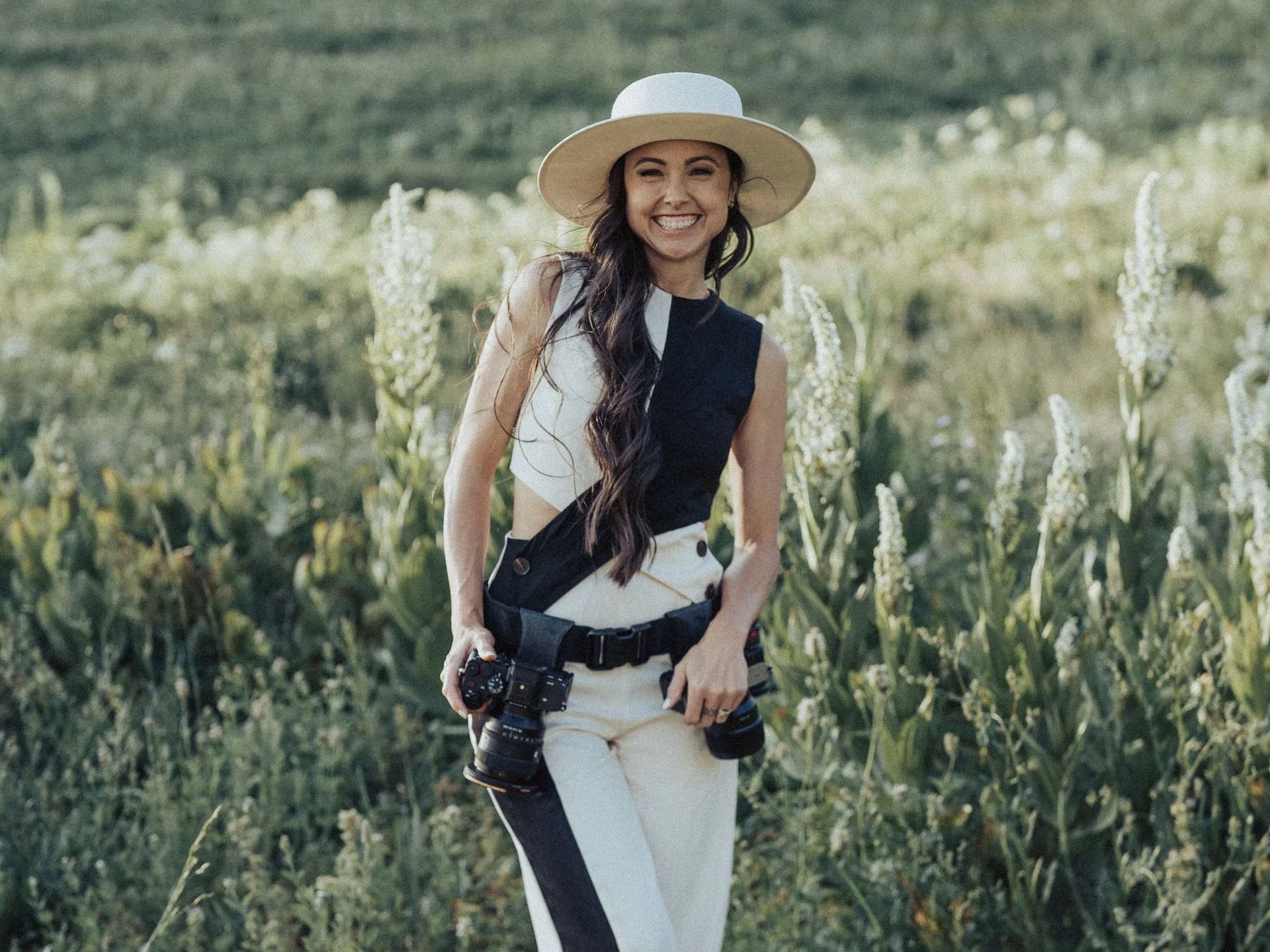 Woman in a white hat and outfit holding a camera, standing in a field of tall green plants.