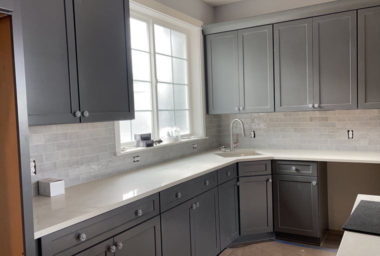 A kitchen with gray cabinets , white counter tops , a sink and a window.