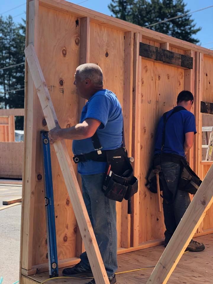 A man in a blue shirt is holding a piece of wood in front of a wall.