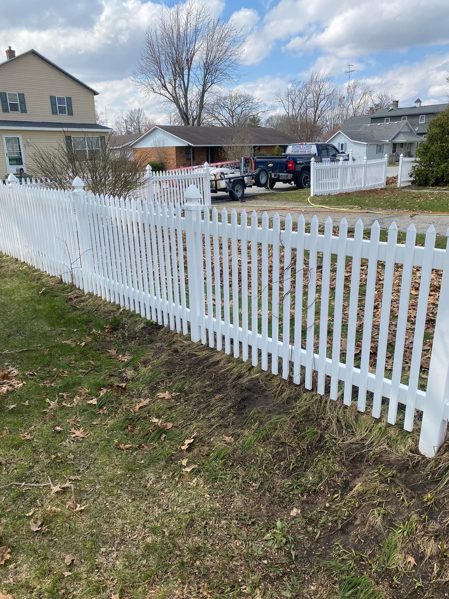 White picket fence in front of a house, curving in the middle, with a truck and car in the background on a sunny day.