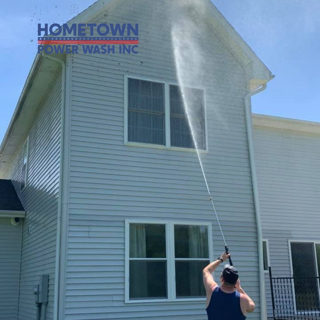 Man pressure washing the side of a two-story house with blue siding on a sunny day.