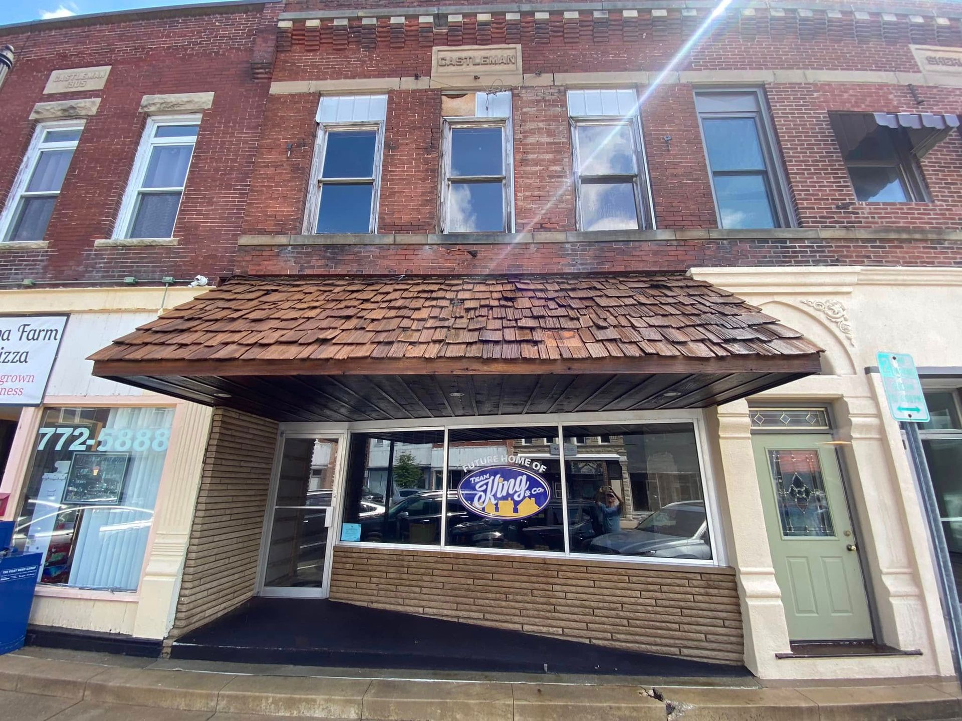 Brick building with a shopfront under a wood-shingled awning; blue sign in the window.
