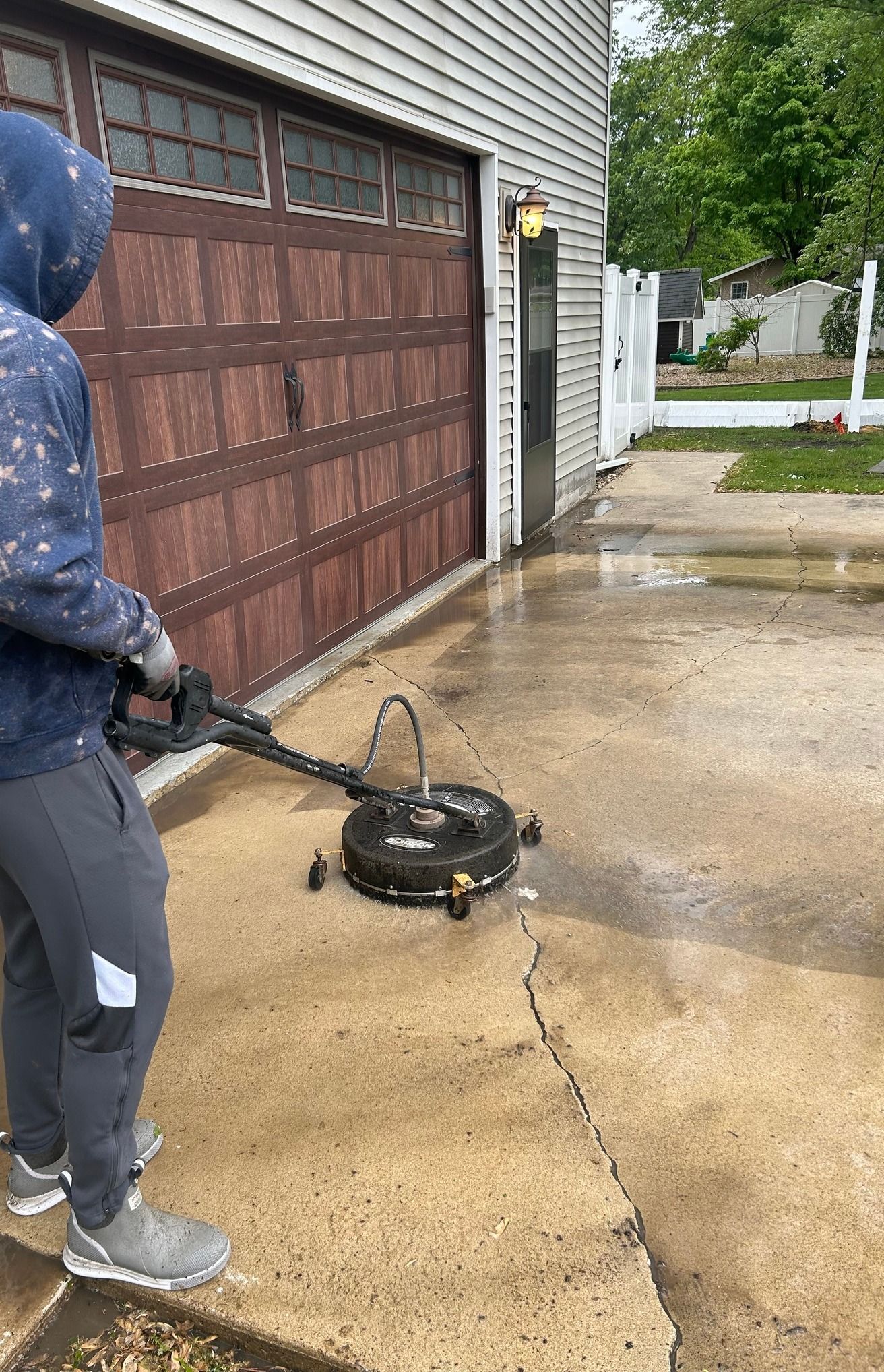 Person power washing a concrete driveway with a surface cleaner next to a garage door.
