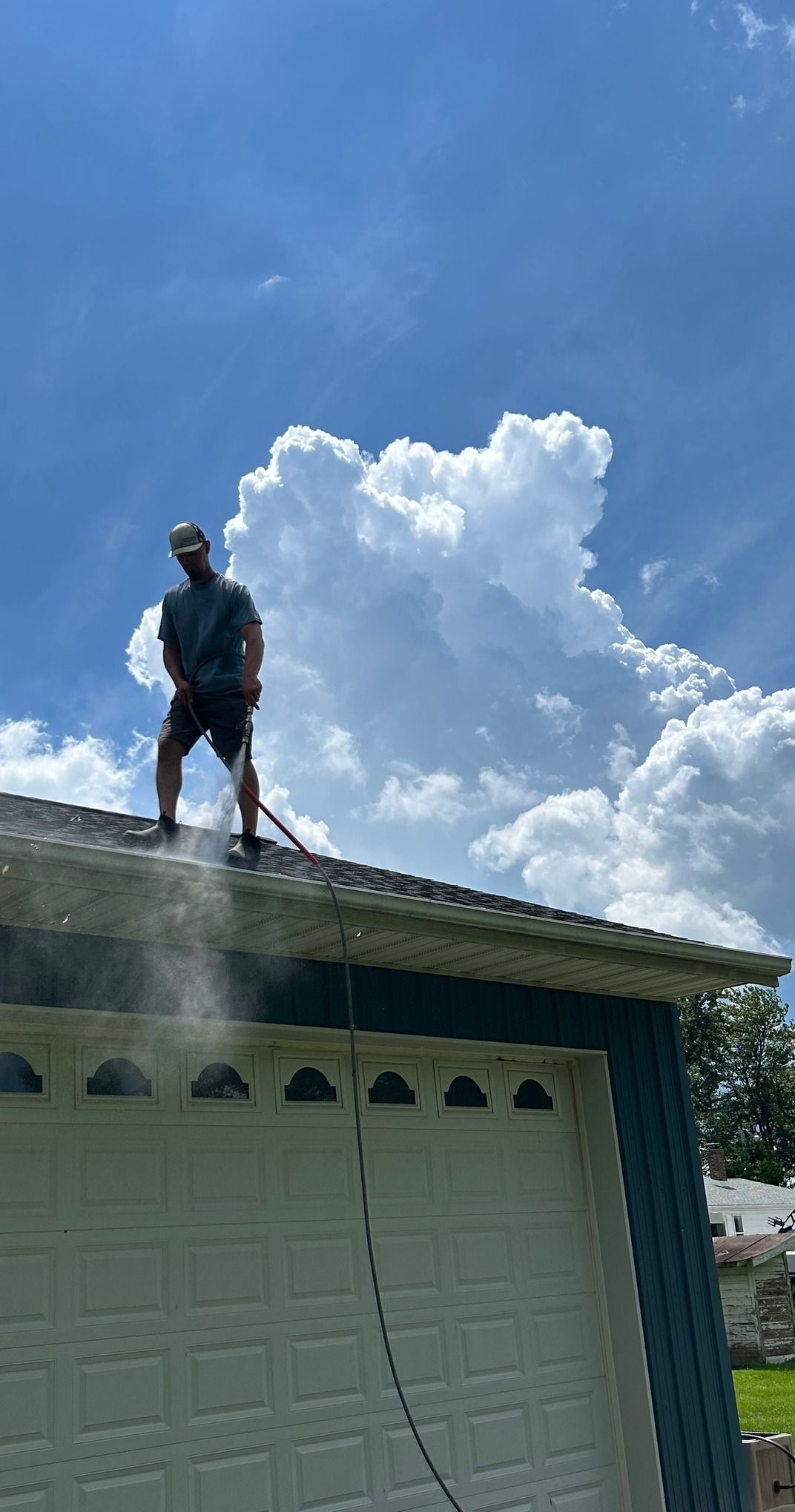 Man pressure washing a roof; sunny day with puffy clouds.