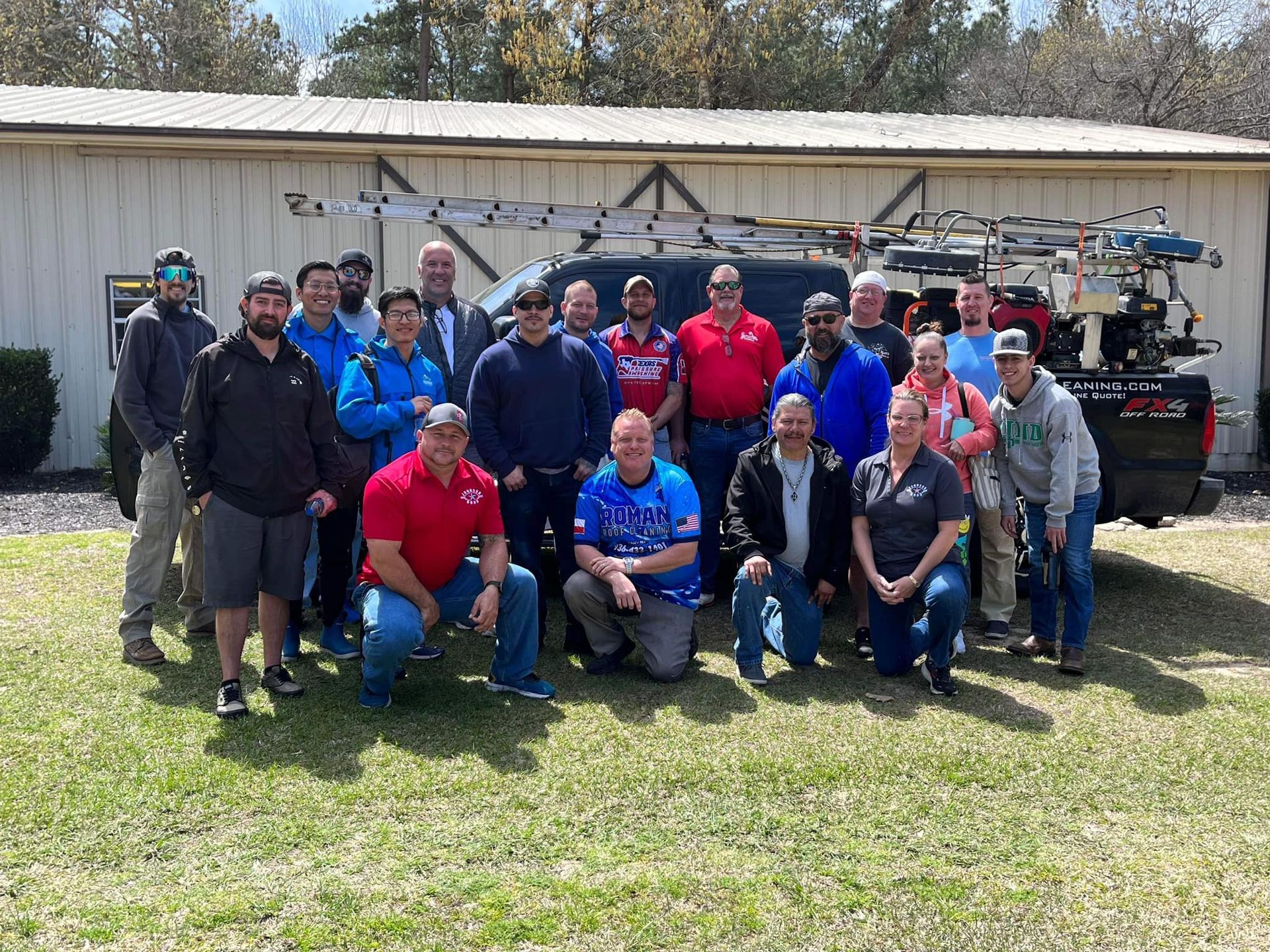 A large group of people standing outside on a grassy lawn in front of a work truck parked near a warehouse building.