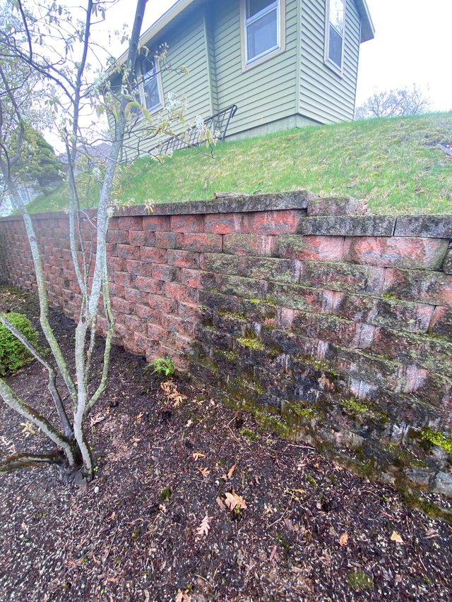 A reddish-brown stone retaining wall sits beneath a green-sided house on a grass-covered slope with a small tree in front.