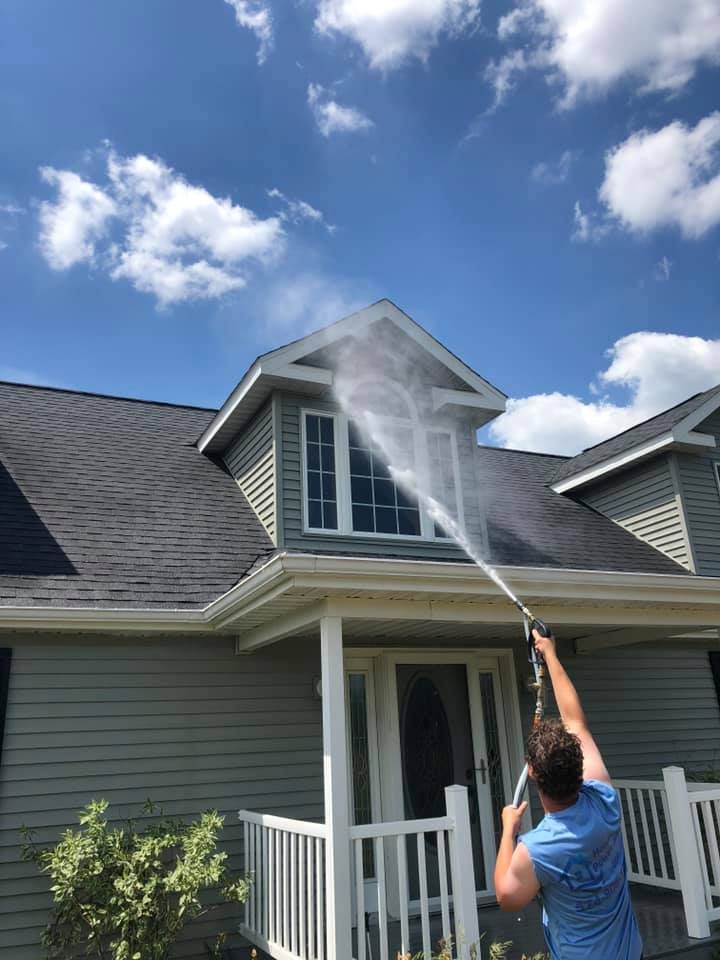 A person in a blue shirt uses a pressure washer to clean the second-story window of a grey house.
