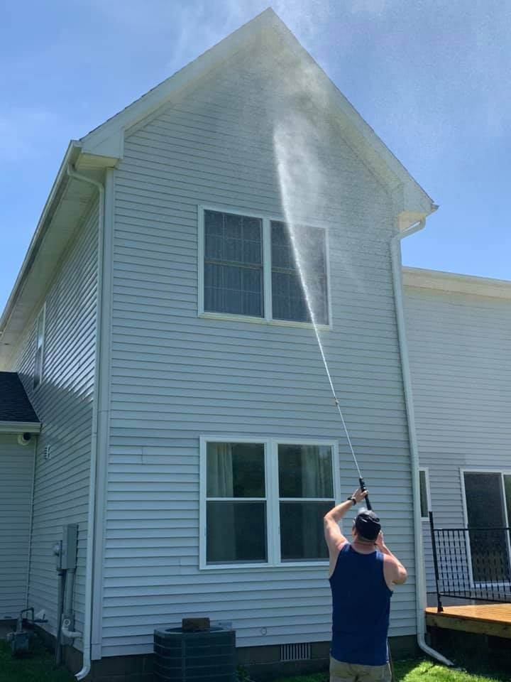 A person using a pressure washer to clean the blue siding of a two-story house on a sunny day.