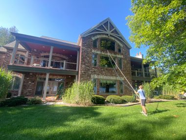 A person uses a long pole to clean the windows of a large, multi-story brick house on a sunny day.