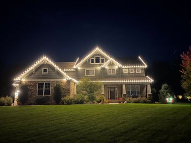 A two-story home at night, illuminated by white roofline holiday lights against a dark sky and green lawn.