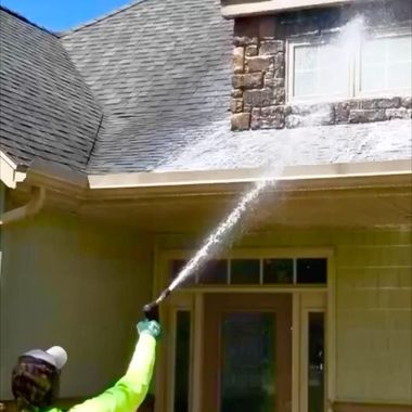 A person in a bright yellow shirt uses a pressure washer to clean the roof of a house with stone accents.