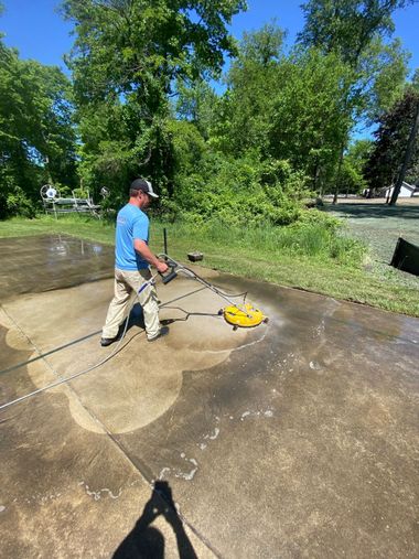 A person in a blue shirt uses a yellow rotary surface cleaner to power wash a dirty concrete driveway outdoors.