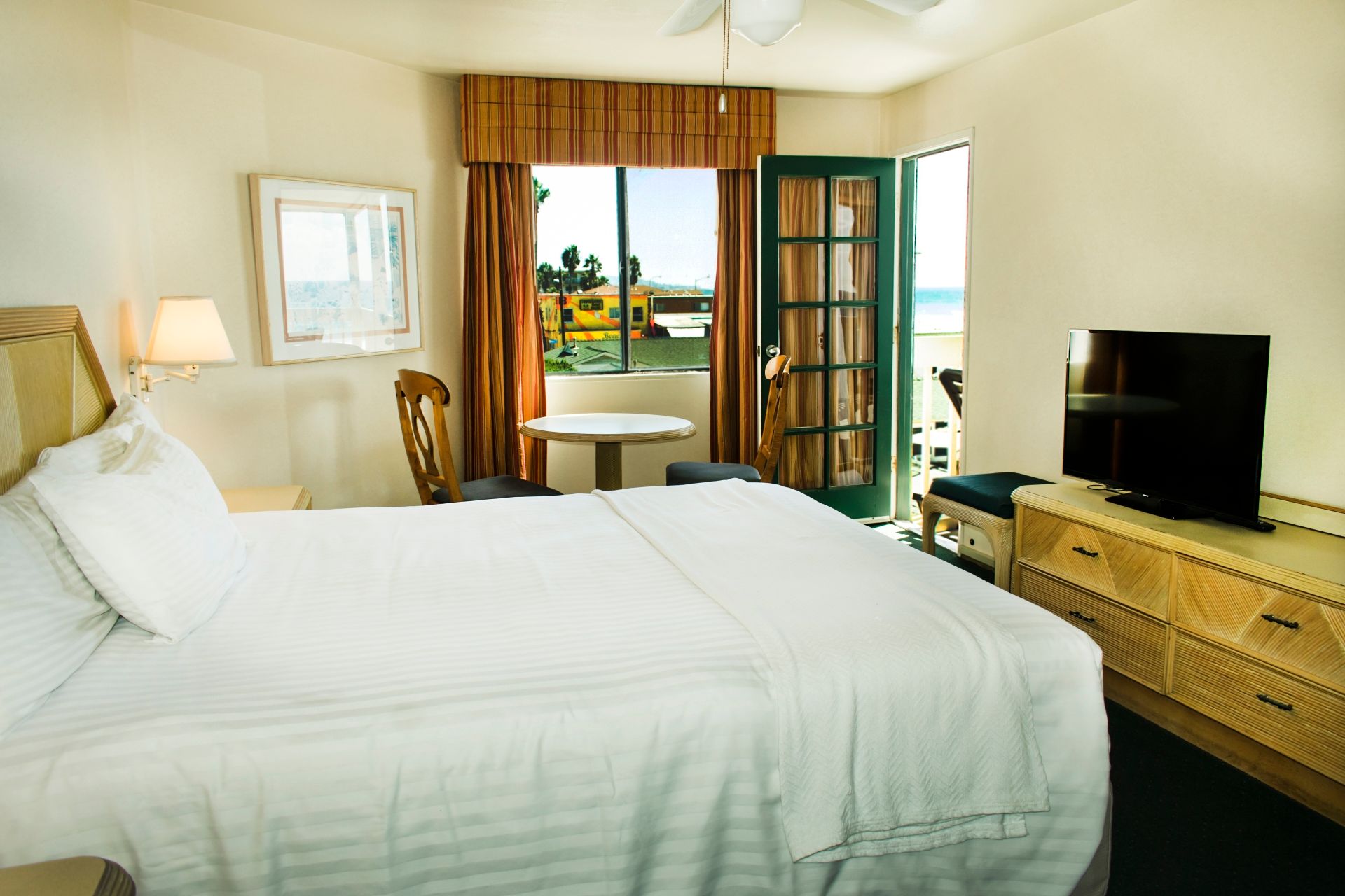 Hotel room with bed, dresser, table, and a door to a balcony overlooking the beach.