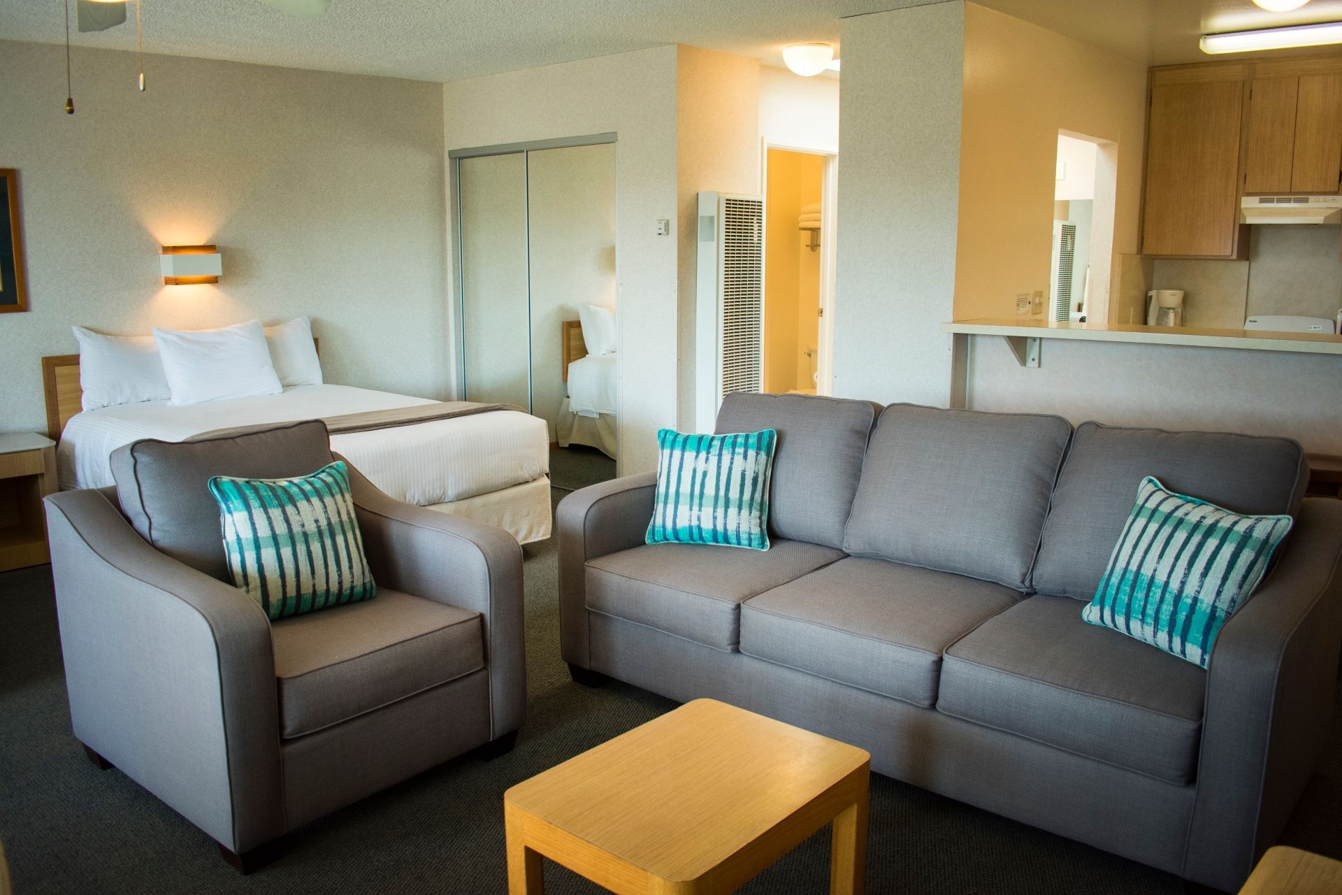 Living area of a hotel room with a couch, armchair, bed, and small kitchen.