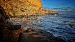 Rocky coastline with waves crashing against shore, cliffside in the background, lit by sunlight.