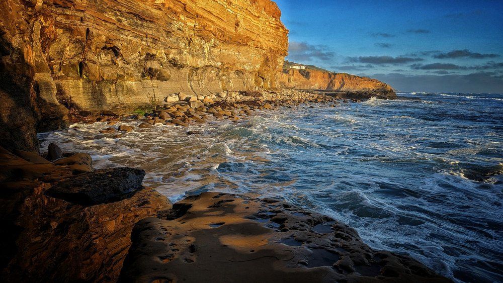 Rocky coastline with waves crashing against shore, cliffside in the background, lit by sunlight.