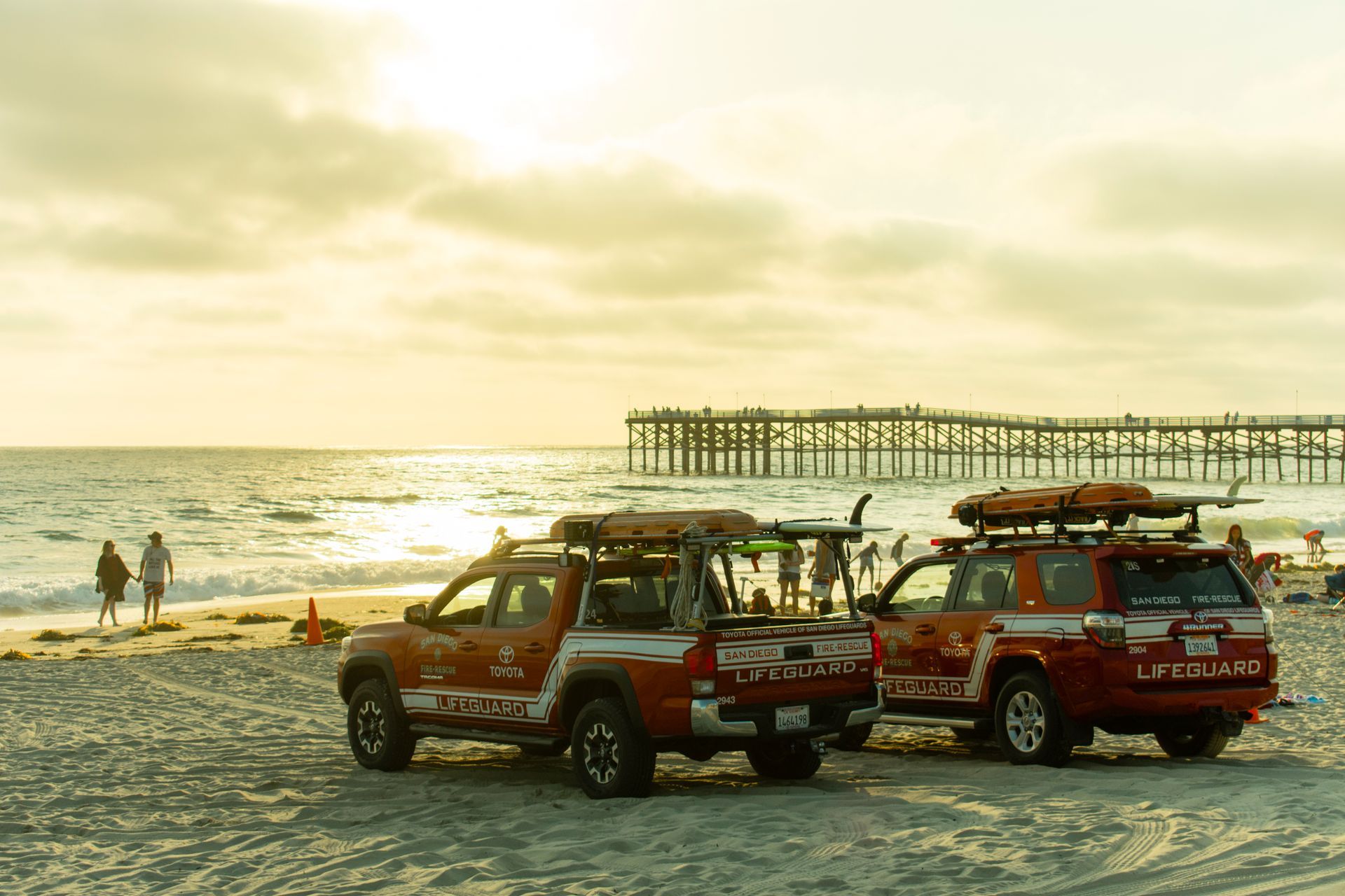 Lifeguard trucks parked on a beach near a pier under a partly cloudy sunsetting sky.