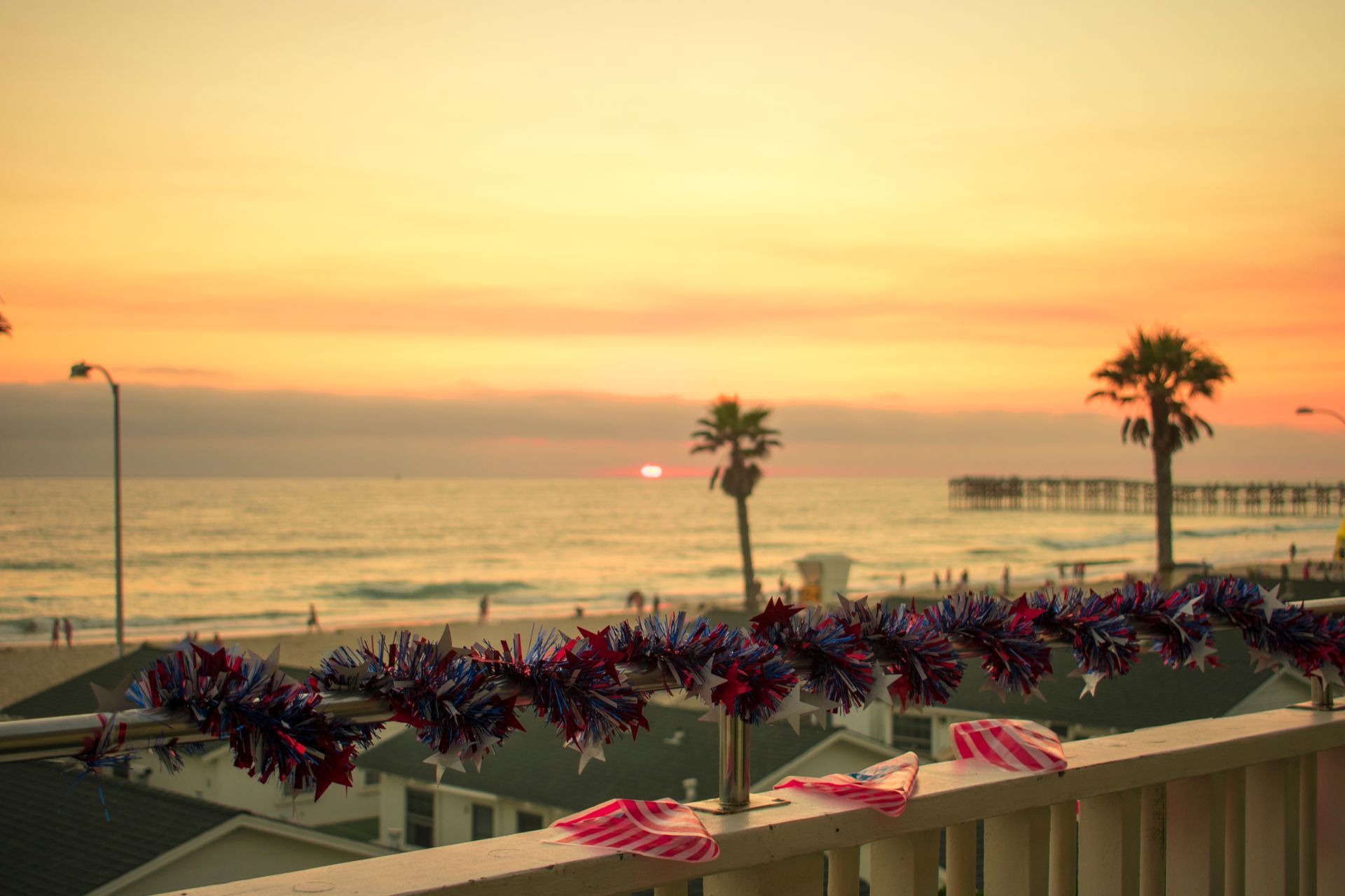 Sunset over beach with palm trees and pier, decorated with red, white, and blue tinsel.