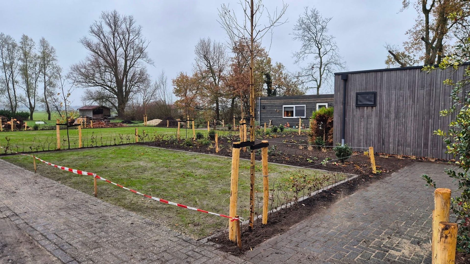 Een grindpad leidt naar een pas aangelegde tuin met jonge bomen, ondersteund door houten palen.
