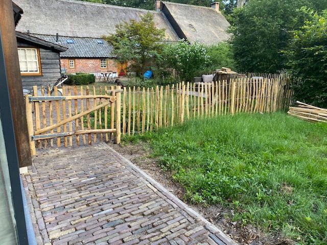 Een houten schapenhek omringt een stenen loopbrug die naar een huis leidt.