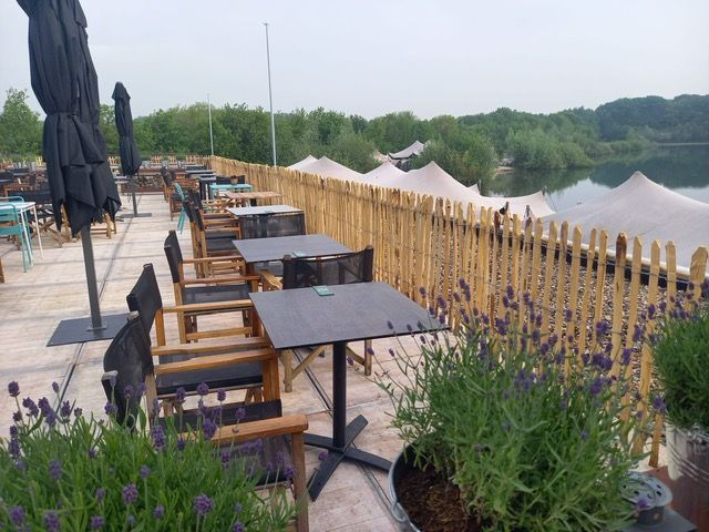 Een terras met tafels, stoelen en parasols met uitzicht op een meer en kastanje hekwerk.