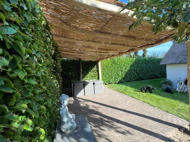 Een hond ligt onder een rieten dak van robinia palen in een tuin.