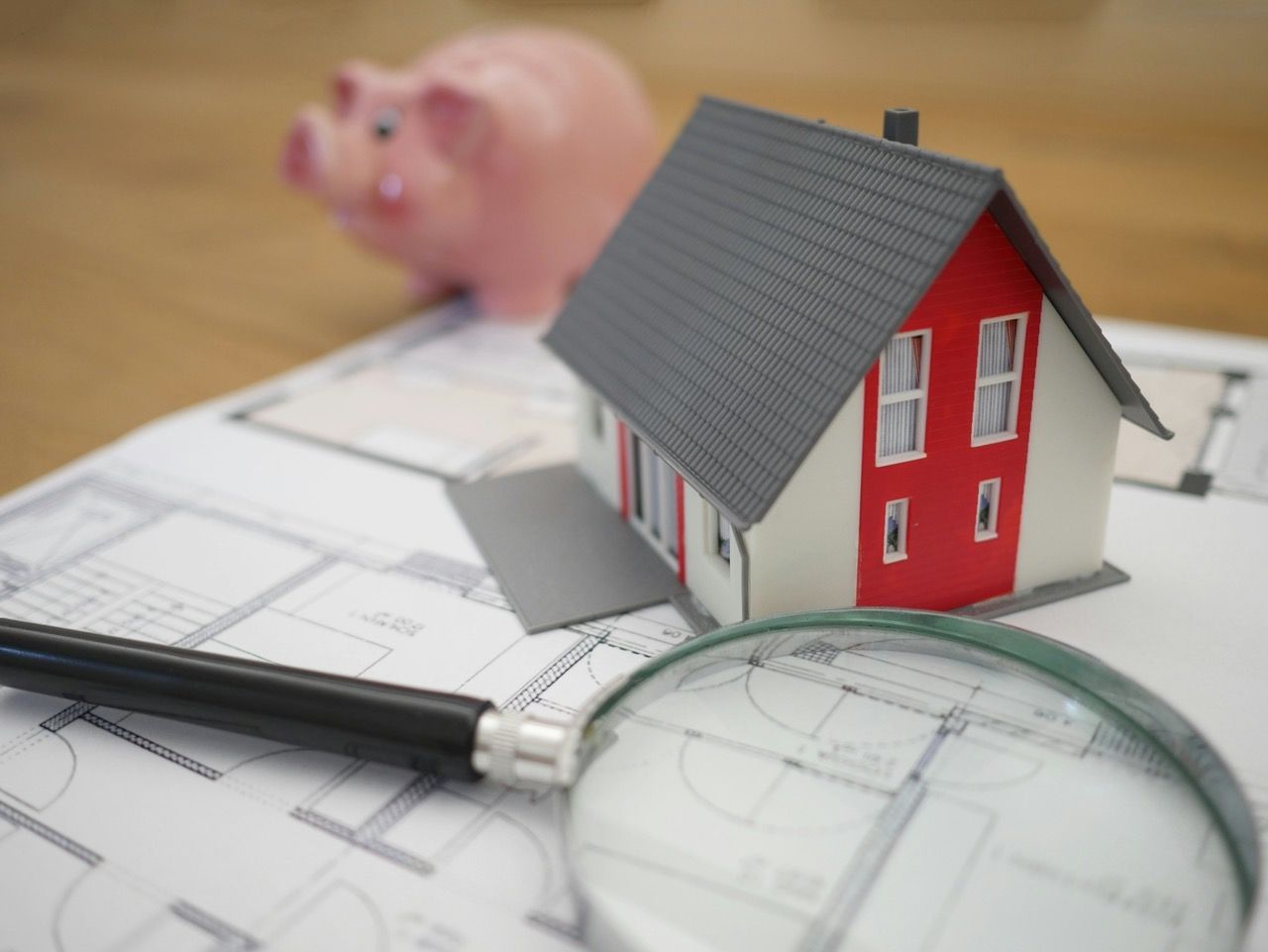 A red miniature house model is placed atop house blueprints with a black pen and a magnifying glass.