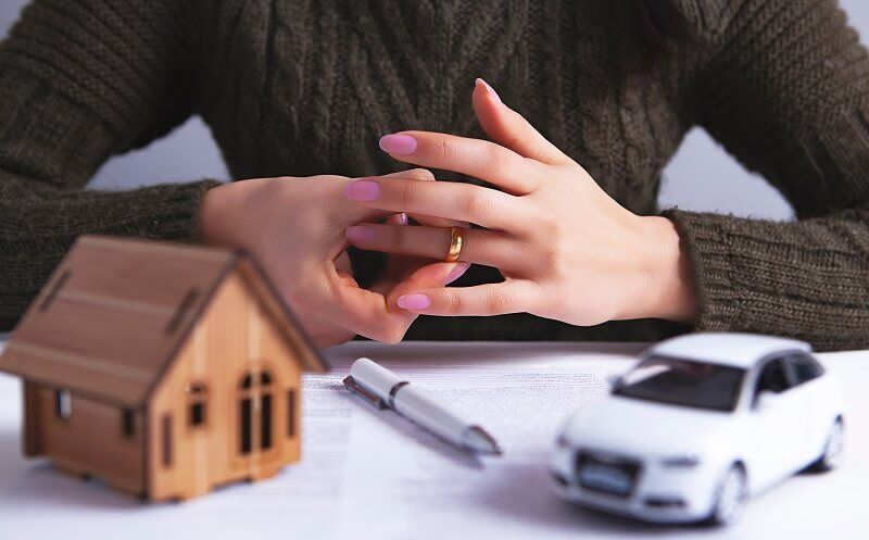 Woman removing wedding ring, with miniature house and legal documents symbolizing divorce.