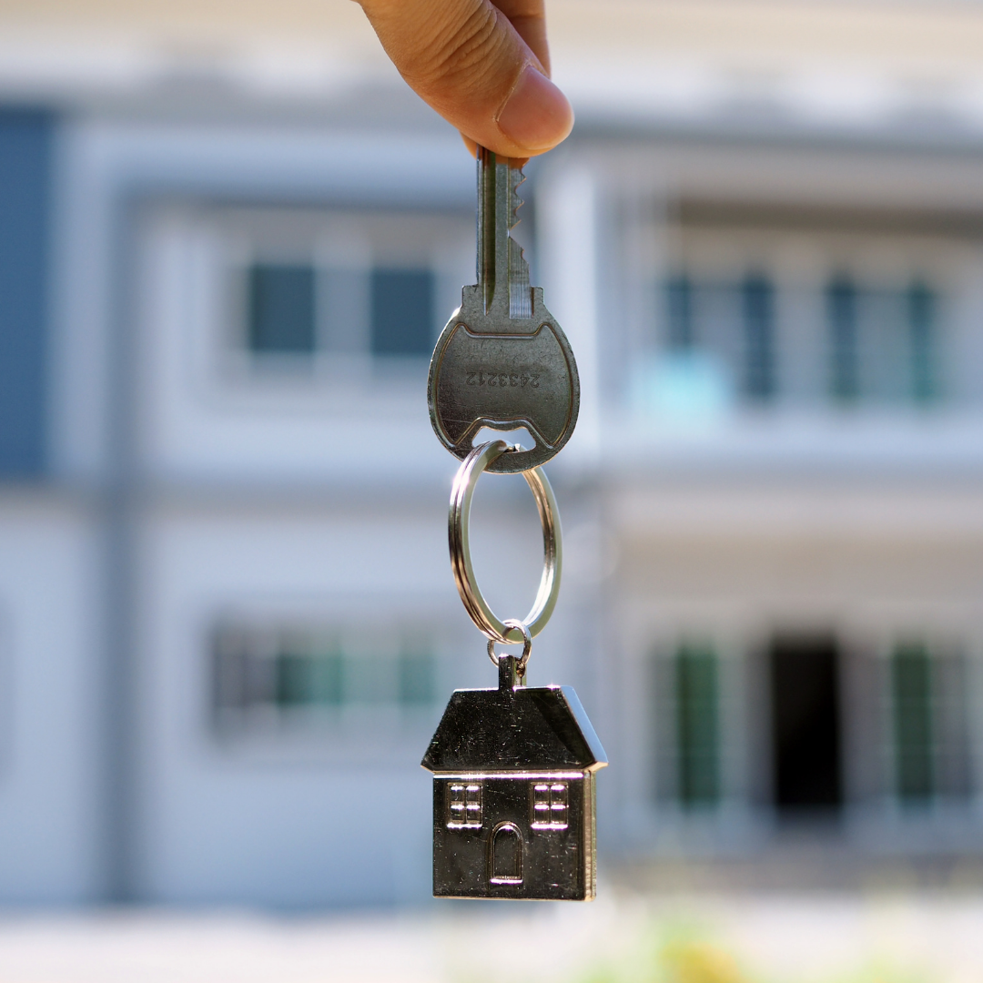 A hand holding a key with a house-shaped keychain, with a blurred building in the background.