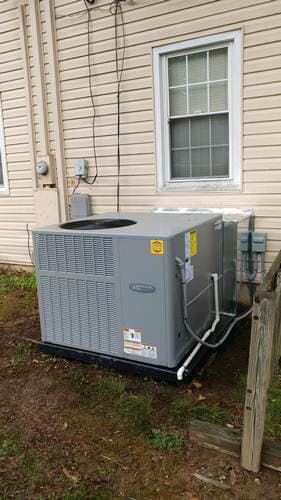 Outdoor air conditioning unit next to a light-colored house with a window, located on a grassy area.