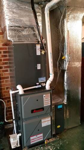 Furnace unit with silver ductwork and white PVC pipes against a brick wall.