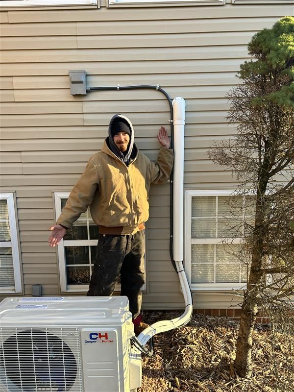 Man stands next to air conditioning unit, pointing up at conduit running along siding.