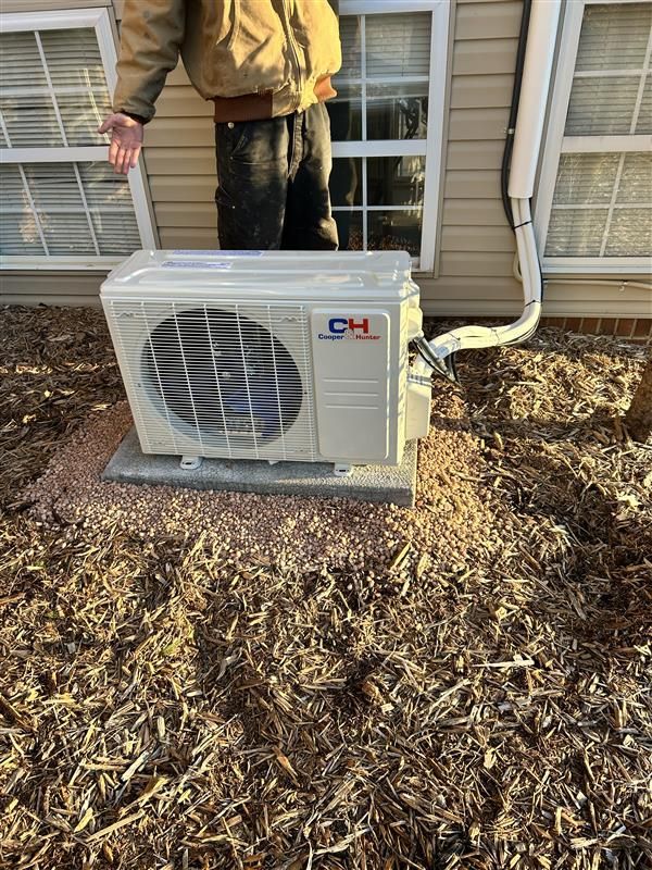 An air conditioning unit is outside a house. A person stands nearby, gesturing with their hand.