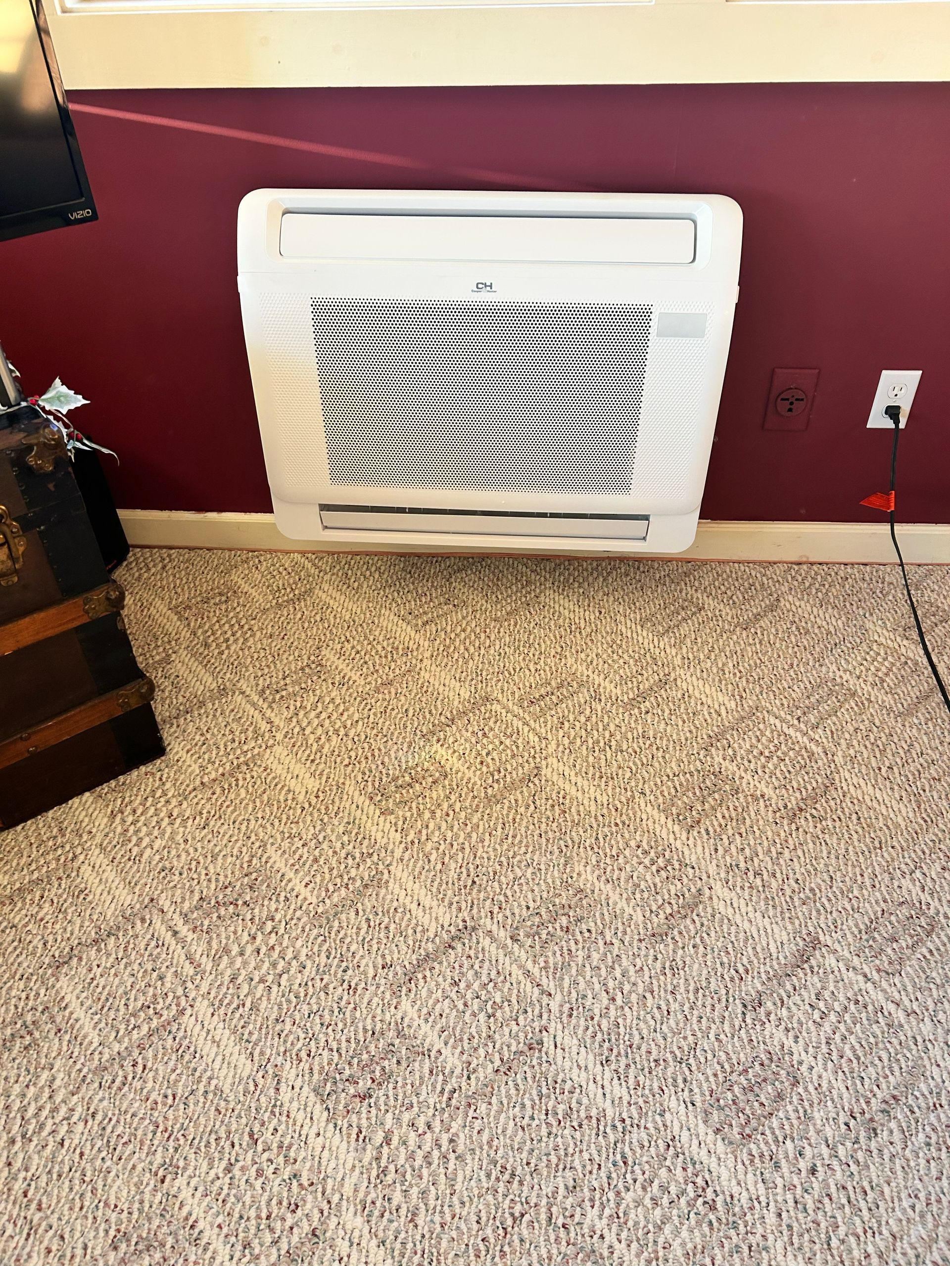White wall air conditioner on a red wall, above a carpeted floor. A trunk and power outlet are also visible.