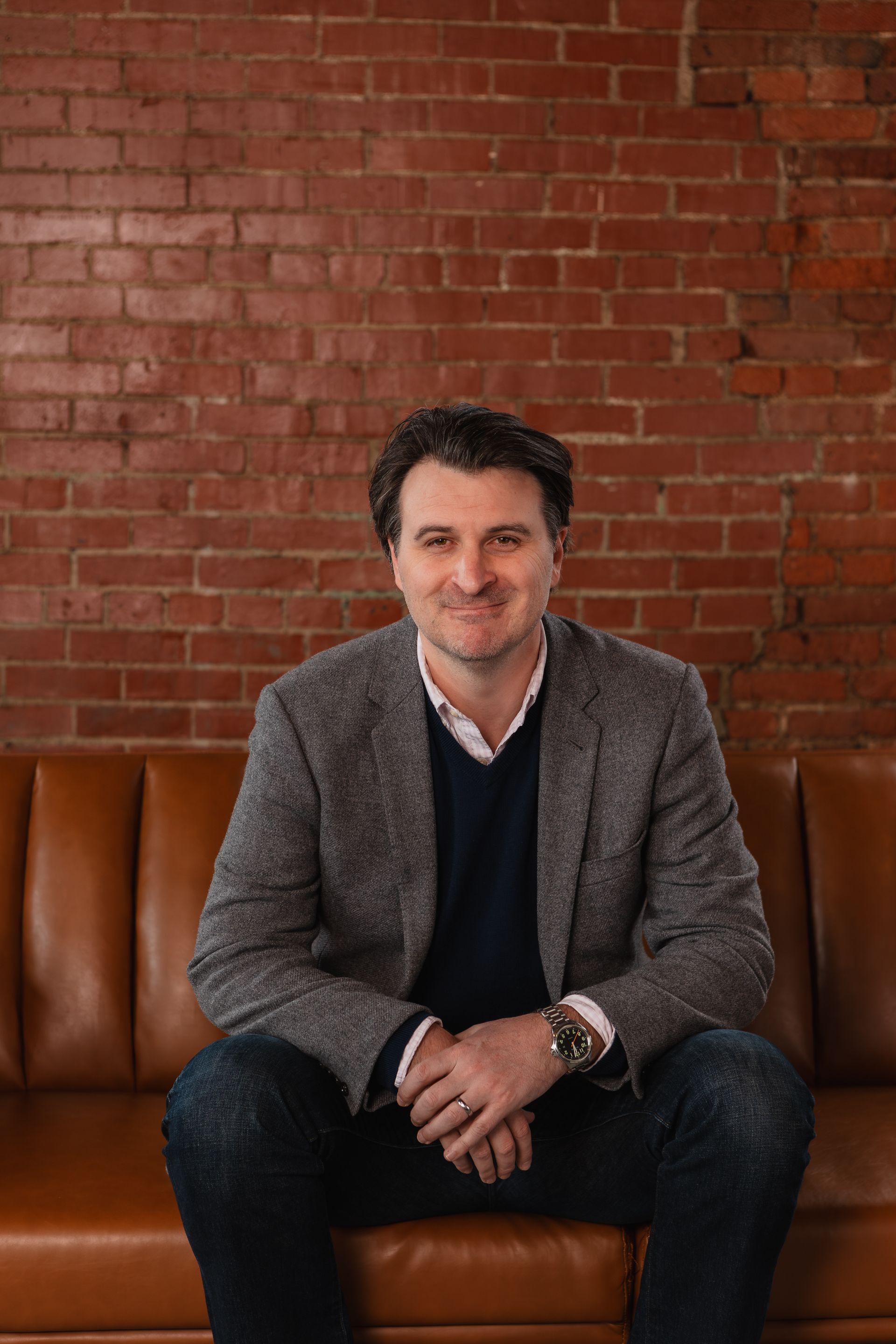 A man in a suit is sitting on a couch in front of a brick wall.