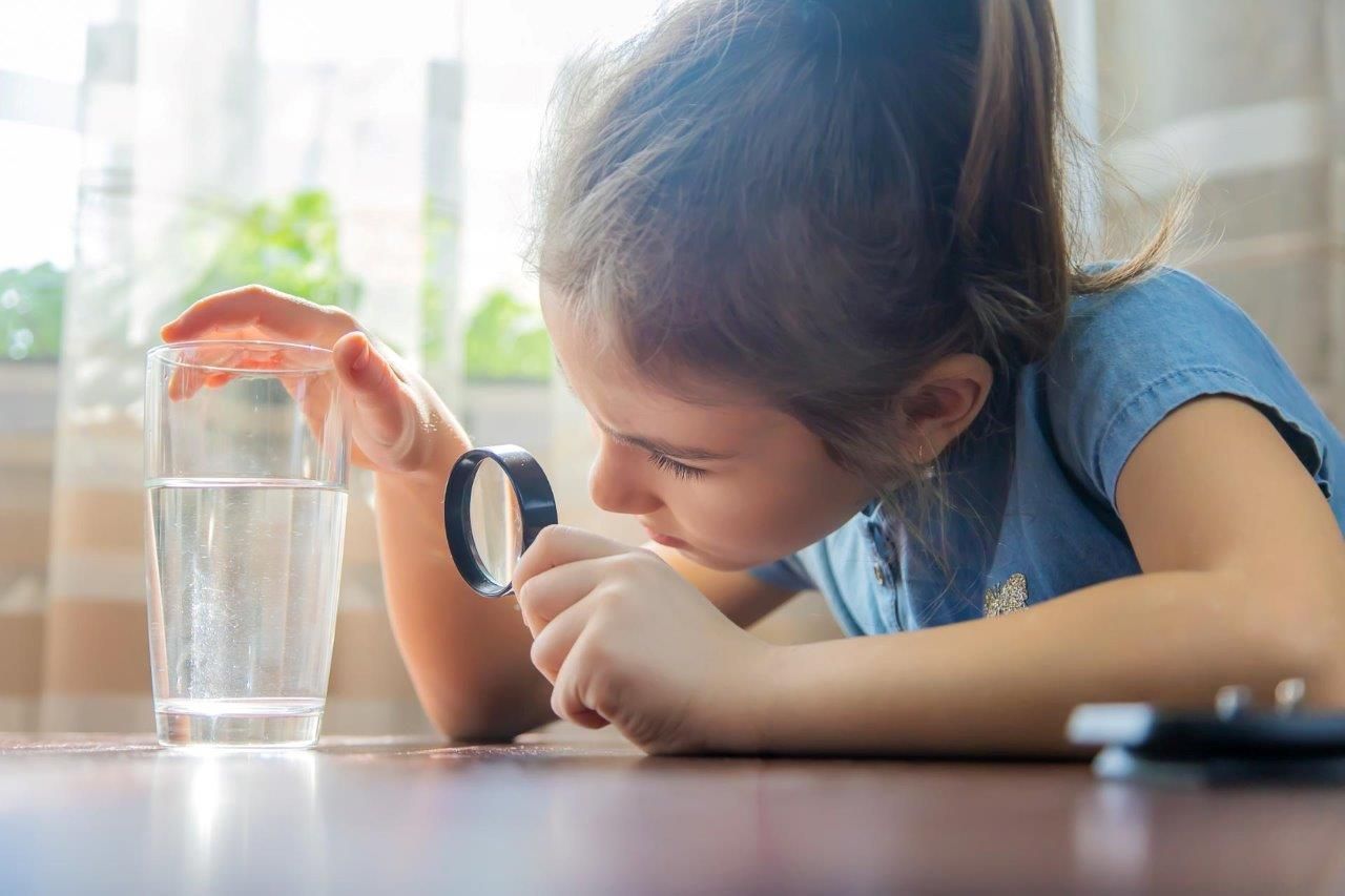 Una bambina guarda un bicchiere d'acqua attraverso una lente d'ingrandimento.